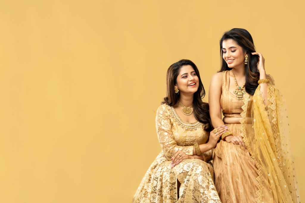 Two smiling women in elegant traditional Indian dresses with a warm backdrop.