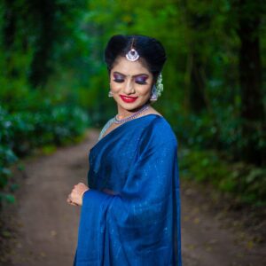 Woman in traditional blue saree standing gracefully amidst a vibrant green forest.